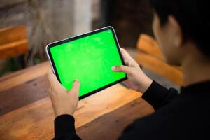 Mock up photo of a close up shot featuring a mans hand holding an iPad tablet with a green screen against the background of a wood cafe table