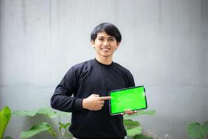 joven asiático hombre con un negro suéter participación y señalando a un ipad tableta verde pantalla Bosquejo foto