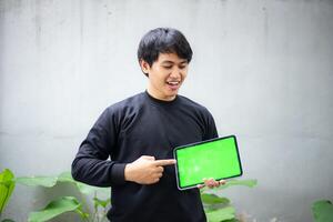 Young Asian man with a black sweater holding and pointing at an iPad tablet green screen mockup photo