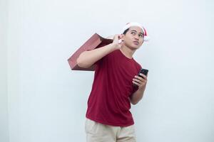 Young Asian man wearing a Santa Claus hat holding a smartphone and a shopping bag with expressions of smile, shock, and surprise, isolated against a white background for visual communication photo