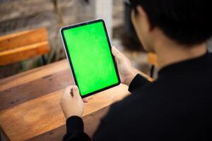 Mock up photo of a close up shot featuring a mans hand holding an iPad tablet with a green screen against the background of a wood cafe table