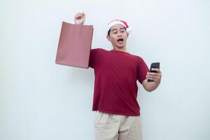 joven asiático hombre vistiendo un Papa Noel claus sombrero participación un teléfono inteligente y un compras bolso con expresiones de sonrisa, choque, y sorpresa, aislado en contra un blanco antecedentes para visual comunicación foto