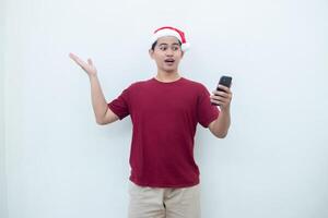 Young Asian man wearing a Santa Claus hat holding a smartphone and expressing a smile, surprise, and pointing to the side, isolated by a white background for visual communication photo