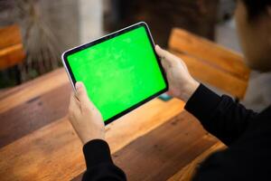 Mock up photo of a close up shot featuring a mans hand holding an iPad tablet with a green screen against the background of a wood cafe table