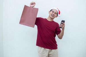 joven asiático hombre vistiendo un Papa Noel claus sombrero participación un teléfono inteligente y un compras bolso con expresiones de sonrisa, choque, y sorpresa, aislado en contra un blanco antecedentes para visual comunicación foto