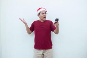 Young Asian man wearing a Santa Claus hat holding a smartphone and expressing a smile, surprise, and pointing to the side, isolated by a white background for visual communication photo