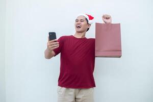 Young Asian man wearing a Santa Claus hat holding a smartphone and a shopping bag with expressions of smile, shock, and surprise, isolated against a white background for visual communication photo