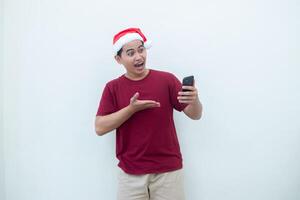 Young Asian man wearing a Santa Claus hat holding a smartphone and expressing a smile, surprise, and pointing to the side, isolated by a white background for visual communication photo