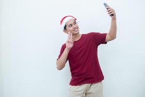 Young Asian man wearing a Santa Claus hat, holding a smartphone and taking a selfie isolated against a white background for visual communication photo