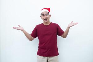 joven asiático hombre vistiendo un Papa Noel claus sombrero sonriente, conmoción y señalando a su lado aislado por blanco antecedentes para visual comunicación foto