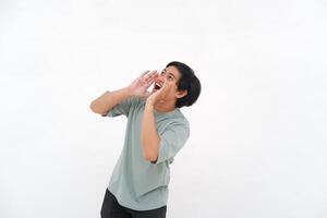 un joven asiático hombre empleado vistiendo camiseta es gritos y gritando ruidoso con un mano en su boca, aislado por blanco antecedentes. foto