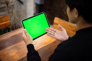 Mock up photo of a close up shot featuring a mans hand holding an iPad tablet with a green screen against the background of a wood cafe table