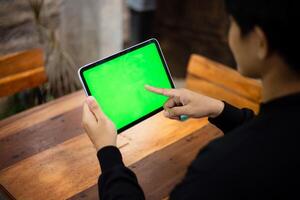 Mock up photo of a close up shot featuring a mans hand holding an iPad tablet with a green screen against the background of a wood cafe table