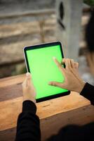 Mock up photo of a close up shot featuring a mans hand holding an iPad tablet with a green screen against the background of a wood cafe table