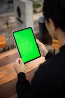 Mock up photo of a close up shot featuring a mans hand holding an iPad tablet with a green screen against the background of a wood cafe table