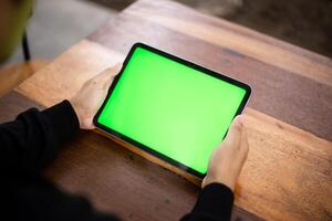 Mock up photo of a close up shot featuring a mans hand holding an iPad tablet with a green screen against the background of a wood cafe table