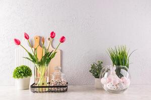 Stylish kitchen background decorated for Easter holiday. vase with tulips, potted plants, eggs on stands, ceramic rabbits. Front view photo