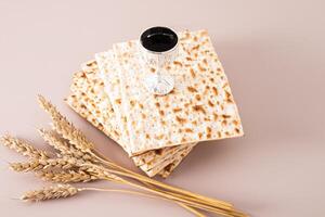 A silver glass of red wine stands on a stack of matzoth slices on a beige background with ears of corn. Concept of the Jewish Passover holiday. photo