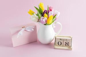 Bouquet of spring tulips in a white jug on a pink background next to a holiday box and a wooden calendar with the date of the eighth of March holiday. photo