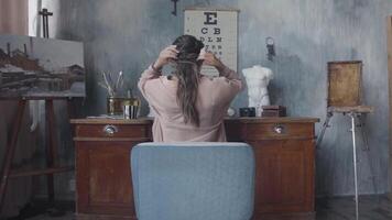 arrière vue de une brunette femme séance à sa bureau et émouvant sa cheveux. Stock images. femme travail dans le art atelier sur gris des murs Contexte. video