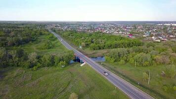 Top view of highway passing through village with forest. Clip. Track with passing cars on background of village with green forest and horizon with sky video