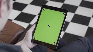 Man in dark jeans sitting on sofa and holding tablet computer with green screen on black and white checkered floor background. Stock footage. Ipad with chroma key. video