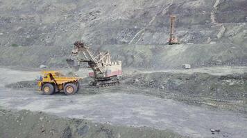 excavatrice et déverser un camion tandis que chargement pierre minerai dans une gris carrière, exploitation minière industrie. action. lourd exploitation minière excavatrice charges Roche minerai dans une benne grand exploitation minière camion, carrière équipement. video