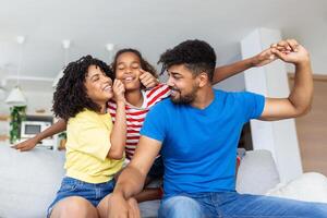 Happy family portrait. Joyful mother, father and their cute daughter posing in living room at home, little girl sitting on dad's shoulders, free space photo