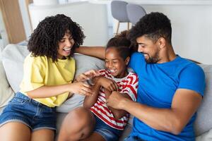 contento multiétnico familia sentado en sofá riendo juntos. alegre padres jugando con su hija a hogar. padre cosquillas su pequeño niña mientras el madre es sonriente. foto