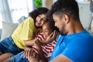 familia imagen de sonriente joven mamá y papá sentar en sofá posando con linda pequeño hija, pequeño emocionado gracioso niña abrazo contento padres, relajarse en sofá juntos foto