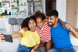 Smiling young family with daughter sit on couch in kitchen make self-portrait picture on cell together, happy parents with small child have fun take selfie on smartphone at home photo