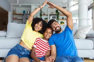 familia cuidado, proteccion y seguro concepto. retrato de sonriente africano americano padres haciendo simbólico techo de manos encima su contento hija, sentado en el piso alfombra en vivo habitación a hogar foto