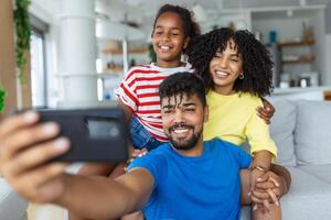 Cheerful family vloggers with child daughter taking selfie, recording vlog, making video call on phone having fun together in living room, parents and kid girl laugh using cellphone funny online app photo