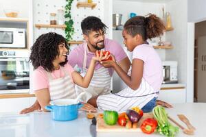 Cute little girl and her beautiful parents are smiling while cooking in kitchen at home. Happy african american family preparing healthy food together in kitchen photo