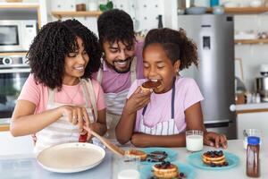 Family At Home Eating Breakfast In Kitchen Together, Happy African American family standing around stove, daughter eating pancakes photo