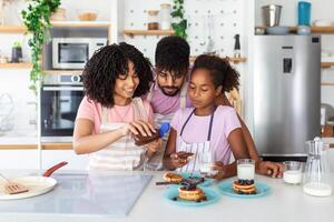 Family At Home Eating Breakfast In Kitchen Together, Happy African American family standing around stove, daughter eating pancakes photo