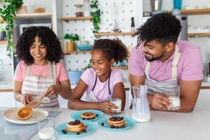 Preparing home bakery. Loving friendly afro american family spouses with daughter cooking food together at kitchen table, making homemade pancakes photo