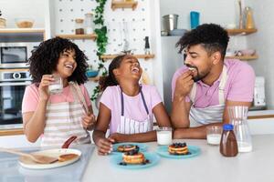 Preparing home bakery. Loving friendly afro american family spouses with daughter cooking food together at kitchen table, making homemade pancakes photo