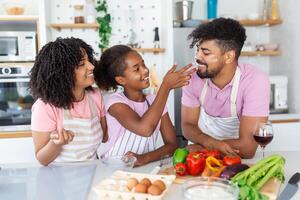 Cute little girl and her beautiful parents are smiling while cooking in kitchen at home. Happy african american family preparing healthy food together in kitchen photo