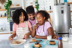 Glad positive young family foolishes with small daughter at kitchen, making pancakes, have tasty breakfast, pose at camera, drink fresh milk, eat pancakes photo