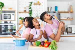 alegre joven padres y pequeño hija teniendo divertido mientras horneando juntos en cocina, contento africano americano familia canto y engañando, utilizando espátula y batidor como micrófonos, Copiar espacio foto