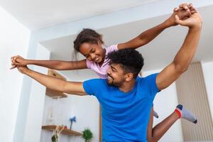 contento padre y niño teniendo divertido jugando adentro. sonriente joven papá y hija gasto hora juntos. contento niña fingiendo a ser un avión foto