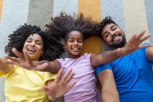 retrato de contento familia posando en piso a hogar, tendido en alfombra y unión, alegremente sonriente, ver arriba. positivo padre, madre y hija caricias, parte superior ver foto