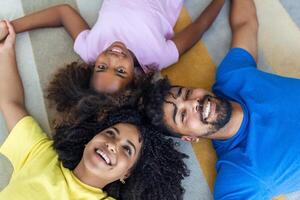 Happy african american family of four bonding lying on bed, black parents and cute little daughter with smiling faces looking at camera in bedroom, mixed race mom dad with child portrait, top view photo