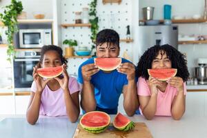 Portrait of cheerful young family taking a bite of a watermelon. African American family standing together at a kitchen eating watermelon. photo