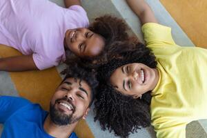 cariñoso africano americano padres y linda hija riendo acostado en piso juntos, contento mezclado carrera familia con niño unión teniendo divertido disfrutar gracioso momentos en dormitorio, parte superior ver desde encima foto