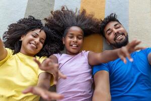 retrato de contento familia posando en piso a hogar, tendido en alfombra y unión, alegremente sonriente, ver arriba. positivo padre, madre y hija caricias, parte superior ver foto