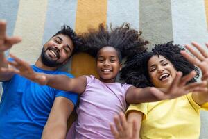 cariñoso africano americano padres y linda hija riendo acostado en piso juntos, contento mezclado carrera familia con niño unión teniendo divertido disfrutar gracioso momentos en dormitorio, parte superior ver desde encima foto