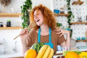 gracioso hermosa mujer canto dentro espátula, Cocinando en moderno cocina, participación espátula como micrófono, baile, escuchando a música, juguetón niña teniendo divertido con batería de cocina, preparando alimento. foto