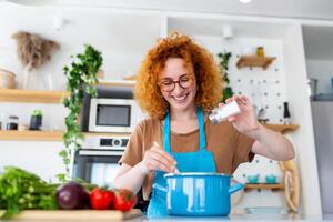 Cute young woman cooking and adding spice to meal, laughing and spending time in the kitchen photo